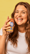 woman smiling and holding a root beer flavor soda leaning against to yellow background